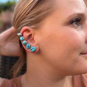 May include: A silver ear cuff featuring four round, bezel-set turquoise stones arranged in a vertical line along the outer edge of the ear. The metal has a polished finish, and each stone displays natural dark matrix patterns within the blue.