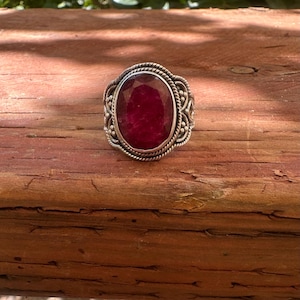 May include: A ring featuring a large, oval-shaped, faceted deep red gemstone set in a silver band. The setting is decorated with intricate twisted rope and small beaded metal details surrounding the central stone, creating a textured border.