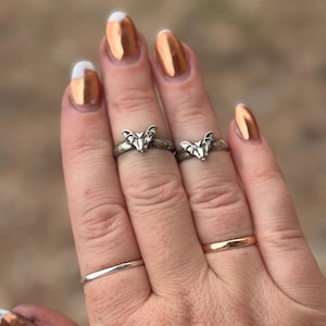May include: Two silver-toned rings featuring a small bat motif on the upper band, paired with two plain, smooth bands worn on the lower fingers. The bat rings have a textured, dark finish, while the plain bands are polished silver and gold-toned.