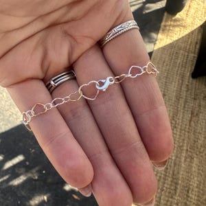 May include: A delicate bracelet featuring heart-shaped links in silver and gold tones. The bracelet is secured with a silver clasp. The jewelry is displayed on a hand, with several rings visible on the fingers.