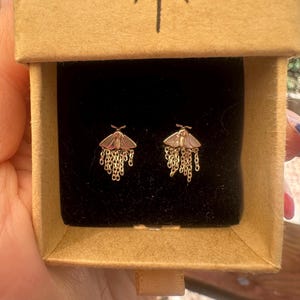 May include: A pair of gold-toned moth stud earrings featuring light pink enamel wings and delicate dangling gold chains. The earrings are presented in a small, square, light brown cardboard box with a black velvet insert and a sunburst icon.