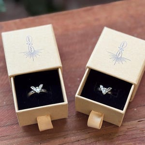 May include: Two silver-toned rings, each featuring a small bat-shaped ornament, are displayed inside individual tan cardboard sliding gift boxes. Each box lid is printed with a blue infinity symbol logo and rests on a black velvet insert.