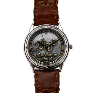 May include: A silver watch with a brown leather strap. The watch face has a silver background with a gold 2000 logo and the text "Walt Disney World".