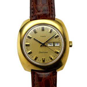 Könnte beinhalten: Eine goldfarbene Timex Electronic Uhr mit einem braunen Lederarmband. Das Zifferblatt der Uhr ist goldfarben mit schwarzen Zeigern und einem Datumsfenster, das "THU 13" anzeigt.