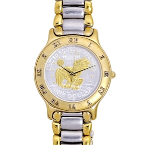 May include: A gold and silver two-tone wristwatch with a silver face featuring a gold American bald eagle and the words "United States of America" and "One Dollar". The watch has Roman numeral hour markers and a two-tone gold and silver band.