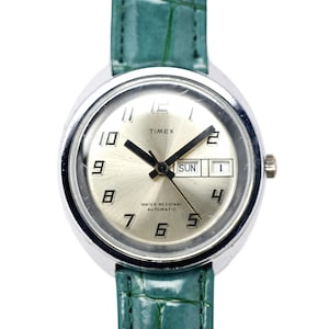 May include: Silver-toned Timex watch with a light-colored face, black hands, and a date display. The watch features a green leather band. The watch face displays the day of the week and the date. The watch is water resistant and automatic.