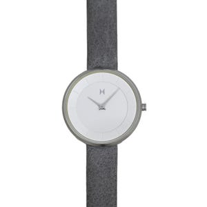 May include: A minimalist wristwatch with a white face and silver-tone case. The watch features a grey suede strap. The watch face has a simple design with silver hands and a logo at the 12 o'clock position.