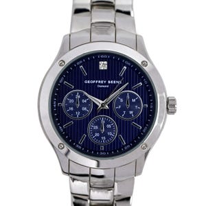 Op de afbeelding: Een zilveren herenhorloge met een blauwe wijzerplaat en drie subwijzerplaten. Het horloge heeft een zilveren band en een diamant op de 12-uur positie. Op de wijzerplaat van het horloge staat "GEOFFREY BEENE Diamond".