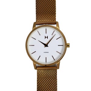 May include: A gold-tone watch with a white face and a mesh strap. The watch face has black hour and minute hands, a black second hand, and a small black 'M' logo at the 12 o'clock position. The watch face also has the word 'AVENUE' printed in black at the 6 o'clock position.