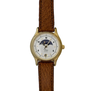 Peut inclure: Une montre Timex Quartz dorée avec un cadran blanc, un design de phase lunaire et un bracelet en cuir marron. La montre affiche la date à 6 heures.
