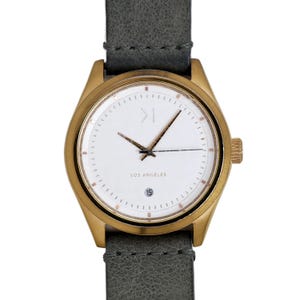 May include: A gold-tone wristwatch with a white face and a grey leather strap. The watch face has a simple design with a small '>' symbol at the top and the words 'Los Angeles' at the bottom.