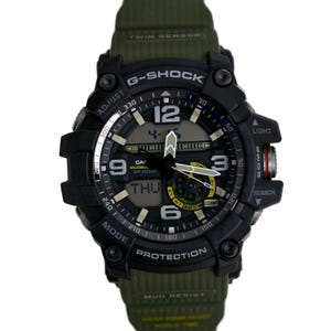 May include: A black G-Shock watch with a green strap. The watch face has a digital display and an analog display with black hands and white markings. The watch has a black bezel with silver markings and a black band with a green woven pattern. The watch has the words "G-Shock", "Mud Resist", "Protection", "Twin Sensor", "Casio", "Mudman", "WR 20BAR", "Comp", "Light", "Search", and "Adjust" printed on it.