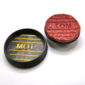 Könnte beinhalten: Zwei schwarze Burger-Pressformen aus Kunststoff mit einem erhabenen Grillmuster. Eine Form hat das Wort "MOT" in goldenen Buchstaben geprägt, die andere das Wort "TOM" in goldenen Buchstaben geprägt.