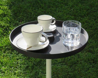 Könnte beinhalten: Ein runder, schwarzer Serviertablett auf einem weißen Ständer, mit zwei weißen Teetassen mit Untertassen, einer kleinen Zuckerdose, einem Löffel und einem Glas Wasser. Die Teetassen haben einen schwarzen Rand. Das Tablett steht auf grünem Gras.