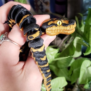May include: A detailed, articulated snake toy in black and yellow. The snake's head is tan with black markings and a yellow eye. The body is segmented with alternating black and yellow sections. The toy is held in a hand.