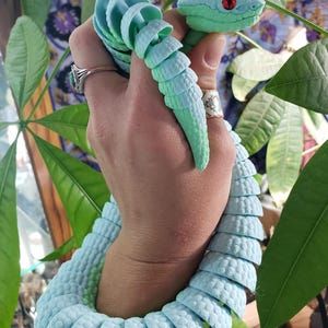 May include: A 3D printed articulated snake toy featuring a light blue and mint green gradient color scheme with textured scales and prominent red eyes. The segmented body is designed to flex and move, wrapping around the wrist in a coiled position.