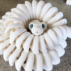 Può includere: Una scultura di serpente bianco arrotolato, con una superficie strutturata e dettagliata. La testa del serpente presenta occhi blu brillante e una bocca dettagliata e aperta. Un'opera d'arte unica.