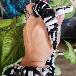 Può includere: Un set di bracciale e anello a forma di serpente articolato in bianco e nero. La testa del serpente ha occhi blu. Il bracciale e l'anello sono realizzati con pezzi segmentati, creando un design flessibile. I gioielli sono tenuti davanti a uno sfondo di foglie verdi.