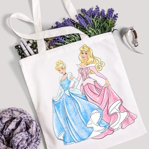 Pu&ograve; includere: Una borsa tote bianca con un designo di Cenerentola e Aurora, le principesse di Cenerentola e La Bella Addormentata nel Bosco della Disney, che indossano i loro iconici abiti da ballo. Le principesse sono una accanto all'altra, Cenerentola a sinistra e Aurora a destra.