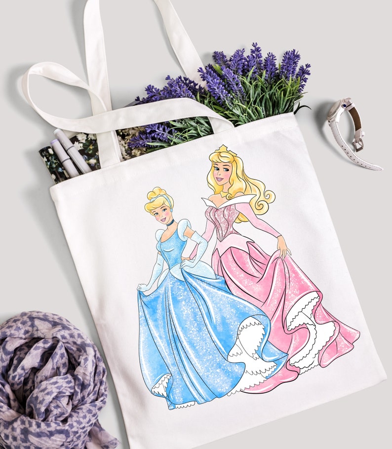 Pu&ograve; includere: Una borsa tote bianca con un designo di Cenerentola e Aurora, le principesse di Cenerentola e La Bella Addormentata nel Bosco della Disney, che indossano i loro iconici abiti da ballo. Le principesse sono una accanto all'altra, Cenerentola a sinistra e Aurora a destra.