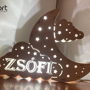 Pode incluir: Uma luz noturna de madeira castanha em forma de lua crescente e nuvem, com recortes de estrelas. O nome "ZSÓFI" é iluminado em branco. A luz está ligada a uma tomada elétrica.