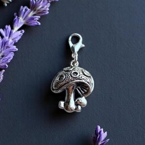 Pode incluir: Um berloque de cogumelo prateado com fecho lagosta. O berloque apresenta um design detalhado com um chapéu e haste texturizados. O fundo é cinza escuro, com raminhos de flores roxas no canto superior esquerdo e inferior direito.