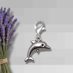 May include: A silver-colored dolphin charm with a lobster clasp. The charm is detailed with fins, a tail, and an eye. The background includes a bouquet of lavender and a gray and white brick pattern.