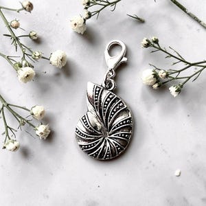 May include: A silver nautilus shell charm with detailed etched patterns and a lobster clasp. The charm is surrounded by sprigs of white flowers and green stems, set against a white background. The charm is approximately 2.5 cm in length.