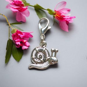 Op de afbeelding: Een zilverkleurige slakkenbedel met een karabijnsluiting, perfect voor een armband of ketting. De bedel is gedetailleerd met een spiraalvormig huisje en antennes. Roze bloemen en groene bladeren zijn op de achtergrond.