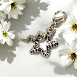 May include: Silver charm featuring a star design with a lightning bolt detail. The charm has a lobster claw clasp.