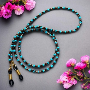 Könnte beinhalten: Brillenkette mit türkisfarbenen und bronzefarbenen Perlen. Die Perlenkette hat goldfarbene Schlaufen zum Halten der Brille. Der Hintergrund ist einfarbig grau mit rosa Blumen in den Ecken.