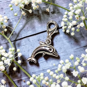 May include: A silver-toned metal dolphin charm featuring a lobster claw clasp. The dolphin is depicted in a leaping position with a smooth, polished surface and a small circular eye detail. The charm is designed for attachment to jewelry or accessories.