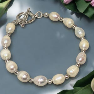 May include: A white pearl bracelet with a silver toggle clasp. The pearls are oval shaped and have a slight iridescent sheen.