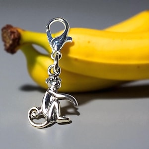 May include: A silver-colored monkey charm with a lobster clasp. The charm is in the foreground, with a yellow banana in the background. The monkey is sitting with its arms outstretched. The charm is likely for a bracelet or necklace.