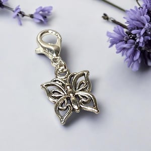 Może przedstawiać: Srebrny charms w kształcie motyla z ażurowym wzorem, przymocowany do karabińczyka. Charms ma szczegółowe linie i krzywizny, sugerujące ręczne wykonanie. Gałązki lawendy stanowią miękkie tło.