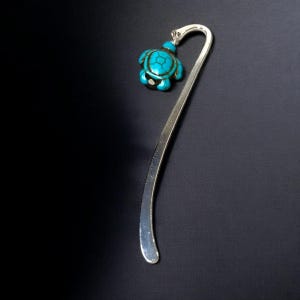 May include: Silver bookmark with a turquoise turtle charm. The bookmark is curved and has a pointed tip.