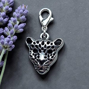 Może przedstawiać: Srebrzysty metalowy charms w kształcie głowy lamparta ze szczegółowymi czarnymi plamkami i rysami twarzy, przymocowany do małego zapięcia typu karabińczyk, umieszczony obok gałązki fioletowych kwiatów lawendy.