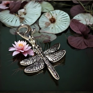 May include: Silver dragonfly charm with a detailed design, hanging from a silver clasp. The charm is resting on a dark surface with dried flowers.