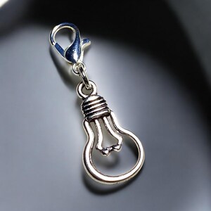May include: A silver-colored charm in the shape of a lightbulb, with a lobster clasp at the top. The lightbulb charm has a detailed filament design and a screw-in base. The charm is likely made of metal and is suitable for jewelry or craft projects.