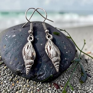Peut inclure: Une paire de boucles d'oreilles pendantes argentées avec un pendentif en forme de coquillage texturé et détaillé. Chaque boucle est suspendue à un crochet métallique simple, présentant un design de coquille en spirale avec des motifs.