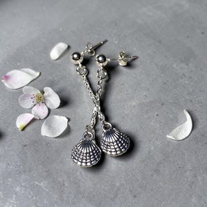 Peut inclure: Une paire de boucles d'oreilles pendantes en argent en forme de coquillage. Chaque boucle d'oreille comporte un petit charme de coquillage détaillé suspendu à une chaîne. Les boucles d'oreilles sont posées sur un fond gris avec des pétales de fleurs blanches éparpillés.