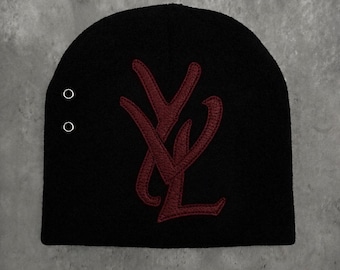May include: A black knit beanie featuring a large, dark red embroidered monogram logo of the letters YL centered on the front. The beanie includes two small, circular silver-toned metal eyelets positioned vertically on the left side panel.