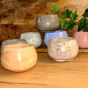 Puede incluir: Seis pequeñas macetas de hormigón en varios colores, incluyendo rosa claro, azul claro, gris y blanco con vetas grises. Las macetas están dispuestas en forma de pirámide sobre una superficie de madera.