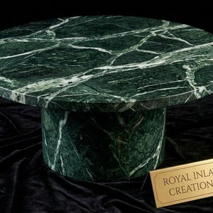 May include: A circular green marble table with white veining. The table has a cylindrical base and a round top. The table is made of polished marble and is set against a black background. The table is a decorative piece. The sign reads "ROYAL INLAY CREATIONS".