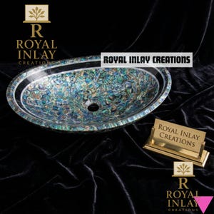 May include: Oval-shaped sink with a mosaic-like pattern of iridescent blue, green, and silver. The sink has a black border and a central drain hole. The text "ROYAL INLAY CREATIONS" is displayed. A gold-colored business card holder is also present.