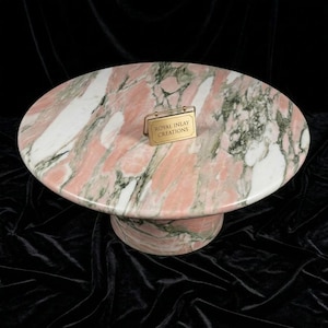 Puede incluir: Mesa ovalada de mármol con un patrón rosa, blanco y verde. La mesa tiene una base cilíndrica y una etiqueta dorada que dice "Royal Inlay Creations."