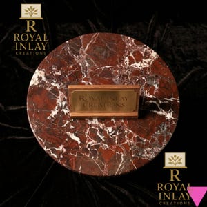 May include: A round, red marble serving board with white veining. A rectangular wooden plaque with the text "ROYAL INLAY CREATIONS" in gold lettering is centered on the board. The background is black.