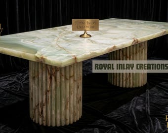 May include: A rectangular onyx table with a green and brown marbled surface. The table rests on two cylindrical bases with vertical ribbed detailing. The text "Royal Inlay Creations" is displayed on the table and in the background.