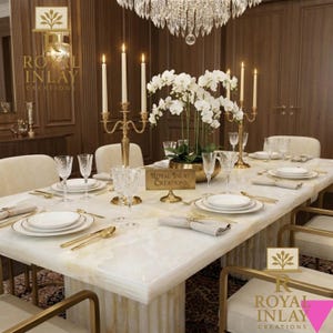 May include: Elegant dining table setting with white plates, gold cutlery, and crystal glassware. Features a centerpiece of white orchids in a gold bowl, lit by candle holders and a chandelier. The table is set for a formal dinner.