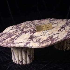 May include: A low coffee table featuring an irregular, organic-shaped top made of cream and purple veined marble. The table is supported by two cylindrical, fluted bases crafted from the same stone. A small gold plaque reads ROYAL INLAY CREATIONS.